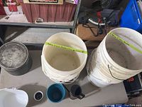 Photo of stacked white 5-gallon buckets, some with drainage holes visible, along with other assorted containers including a metal bucket and smaller plastic vessels.