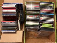 Two boxes filled with about 80 assorted CDs including Christmas music and various artists; some jewel cases and paper sleeves visible; small black CD storage pouch included.