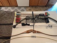 Full layout showing compound bow, arrows, recurve bows, and case on table.