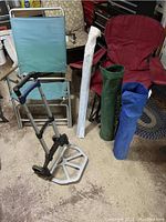 Photo shows four folding chairs: one green lawn chair with wooden armrests and three camping chairs in red, blue, and green. Also visible are three carrying bags (green, blue, white) and part of a collapsible hand truck's base and frame.