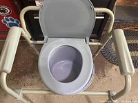 Front view of the commode showing plastic seat with lid open and attached beige metal frame with padded arm handles.