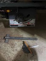 Close-up of saw adjustment control panel with Craftsman 8-inch direct drive label and two metal accessory pieces on the floor