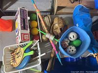 Photo of various outdoor sports items including baseball gloves, croquet mallets, tennis rackets, and a bucket with balls and cones