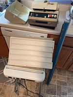 Slide projector on small white table with slide sorter tray and rolled screen leaning next to it.
