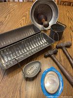 Overall lot contents with seed strainer, two hammers, bread pans, sifter, egg holder and small metal bucket on wooden table.