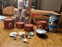 Photo showing the Schmidt's beer box, four Prince Albert tobacco cans, the Whitehead Tankard draught beer tin, several figurines, mini plates, and a small white metal pot with lid arranged on a table.