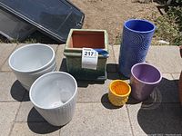 Wide view showing all seven ceramic plant pots with various colors and textures displayed outdoors on a paved surface.