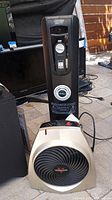 DeLonghi oil-filled heater and Vornado fan positioned outdoors on ground surface, showing front views and power cords.