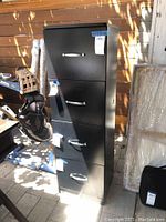 Front view of the black metal filing cabinet with four drawers and silver handles, outdoor setting in natural light.