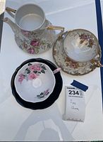Photo showing a coffee pot with pink rose pattern, a teacup with gold floral pattern paired with a saucer, and a teacup with pink rose design and black saucer.