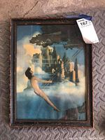 Front view of framed print showing a nude figure swinging with a castle and clouds in the background, some staining and frame wear visible.