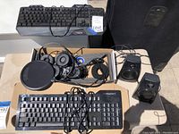 Overview photo showing all keyboards, headphones, mouse, and speakers with tangled cables, taken in outdoor lighting on paved surface.
