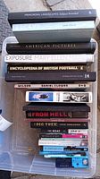 Stack of various hardcover and paperback books shown from top view inside a plastic container.