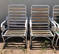 Two weathered iron patio chairs with slatted backs and seats, curved armrests, placed on dirt ground outdoors.