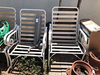 Two iron patio chairs shown outdoors with slatted seats and backs, weathered finish, and decorative curled armrests.