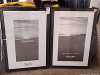 Two large black plastic photo frames side by side, one Aaron Brothers and one Metropolitan, both sized 24 x 36 inches.