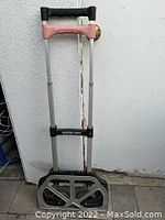 Photo of Magna Cart Personal Folding Hand Truck standing upright against white wall. Shows aluminum frame, black and gray base with wheels, telescoping handle with pink grip sleeve.