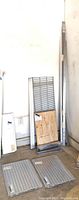 Metal rack parts leaning beside a wall showing shelving panels, plywood board, vertical frame supports and other metal bars unassembled.