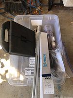 Top-down photo of plastic storage bin containing curtain rods, a Moen towel bar box, black plastic case, and assorted mounting hardware in plastic bags.