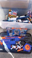 Plastic bin containing N-Strike Elite toy blasters, boxed Explore HD Action Camera, 'Box of Jokes' game, and other small items, plus black lid underneath.