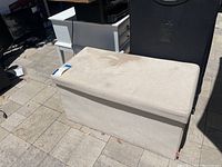 Beige outdoor fabric ottoman with visible dirt and stains, showing hinged lid and rectangular shape.