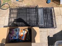 Two black metal wire dog crates with plastic trays placed side by side with a cardboard box of pet care items.