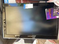 Front view of a black Sceptre flat panel HDTV, 32-inch size, with a blue and white sticker on the top bezel and a product pamphlet showing 32 Sceptre HDTV attached to the bottom right corner.