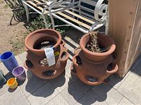Two large terracotta planter pots with multiple side openings placed outdoors next to a bench and building corner.