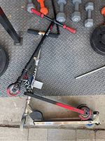 Two scooters on a rubber gym floor with weights in the background. One is a Razor scooter, black and red, the other is a Madd Gear Carve Elite stunt scooter with red foam grips.