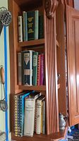 Photo showing wooden shelves filled with assorted antique and vintage cookbooks in various sizes, colors, mostly hardcover, tightly packed next to kitchen utensils on a wall-mounted wooden shelf.