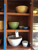 Overall view of five vintage ceramic mixing bowls on wooden shelves, showing yellow, green, white, and blue bowls in various sizes.
