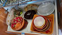 Several ceramic bowls and serving dishes of various colors and sizes arranged in a cabinet drawer. Includes nested bowls primarily in orange, brown, cream, white, and red colors and a brown oval platter.