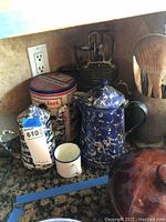 Photo of vintage blue and white marbled enamel coffee pots, white enamel mug with blue rim, Cracker Jack tin, metal egg beater mixer, and wooden utensils container on kitchen counter.