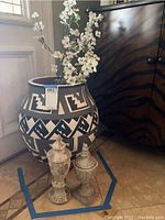Large ceramic pot with white floral arrangement standing next to cabinet, two decorative urns on the floor in front.
