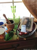 Wide view showing ceramic pig platters, green glass bottle, wooden mortar and pestle, green glass duck, ceramic mugs, and a photo card