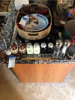 Photo showing multiple sets of vintage salt and pepper shakers of various materials including wood, ceramic, metal, and glass arranged on a countertop, accompanied by a woven basket and decorative plate.