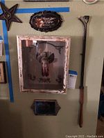 Wall display featuring a vintage angel shadow box with distressed white frame, black oval engraved metal sign above it, smaller rectangular metal plaque below, and a tall black metal candle snuffer leaning against the wall.