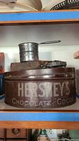 Photo of vintage Hersheys Chocolate & Cocoa metal tin with lid, round shape, brown color with white lettering, shelf top view.