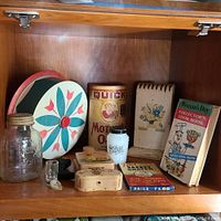 Vintage kitchen items displayed inside wooden cabinet including cake tin, glass jar, cookbooks, recipe pad and other accessories.