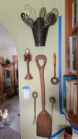 Photo showing the collection of vintage kitchen utensils arranged on a wall including a wooden spatula, metal whisks, and a decorative metal holder.