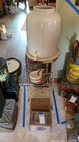 Full view of the vintage ceramic water dispenser with spigot on metal stand with ceramic vessels and wooden box below