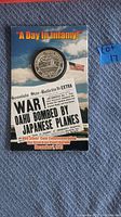 Coin displayed on a blue surface alongside a newspaper headline reading 'WAR! OAHU BOMBED BY JAPANESE PLANES' and other Pearl Harbor themed text.