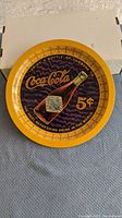 Top view of the round Coca-Cola 75th Anniversary metal tray showing central bottle graphic with 5 cent price and yellow border.