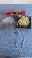 Photo showing the vintage plastic coin bank resembling a large Liberty coin with a red level above, a small blue numbered marker, and the coin bank on a clear plastic stand on a blue surface.