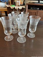 Seven clear parfait glasses with etched diagonal crisscross designs arranged on a dark wooden table.