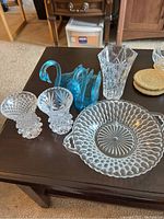 Photo showing all glassware items including two small crystal vases, larger crystal vase, two blue glass swans, and clear serving dish on a brown wooden table.