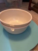 Two white ceramic soup bowls stacked on top of a light blue round ceramic plate, all shown on a carpeted floor.