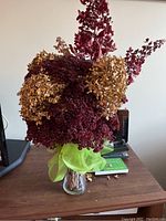 Full view of dried flower arrangement with orange, burgundy and brown flowers and green mesh decoration in clear glass vase on wooden table.