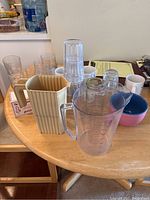 Photo showing assorted glassware, mugs, a beige ribbed ceramic milk jug, small bowl with lid, and various glass and plastic drinking glasses arranged on a oval wooden table.