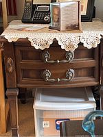 Front view of a wooden side table with drawer, showing decorative handles and a lace table cover, with objects placed on top and beneath the table.